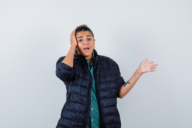 Young woman pointing right, with hand on head in shirt, puffer jacket and looking puzzled. front view.