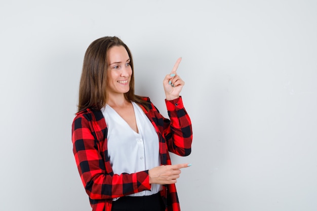 Young woman pointing up and right, looking away in casual clothes and looking cheery. front view.