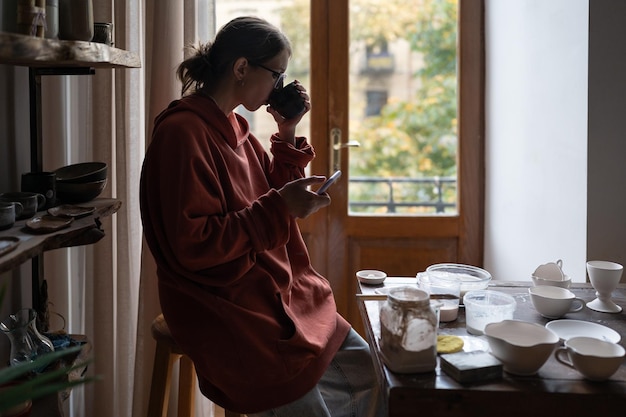 Young woman potter resting in cozy home pottery studio drinking tea and using smartphone