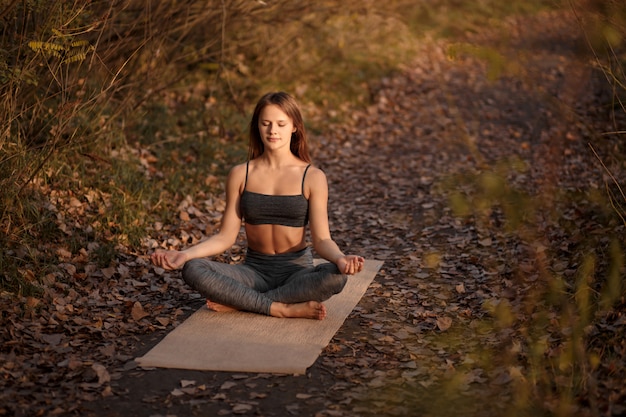 Young woman practicing yoga exercise at autumn park with yellow leaves. Sports and recreation lifestyle