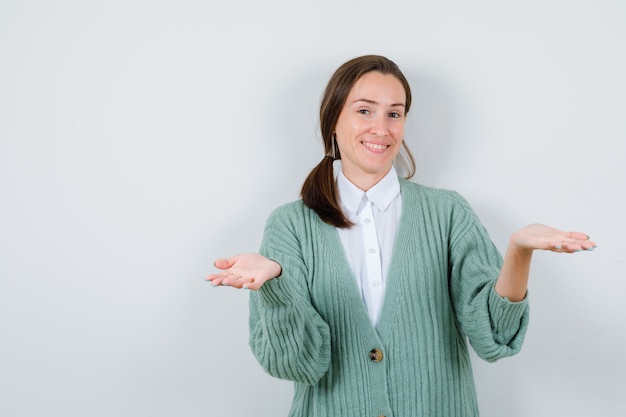 Young woman pretending to offer something in blouse, cardigan and looking cheery , front view.