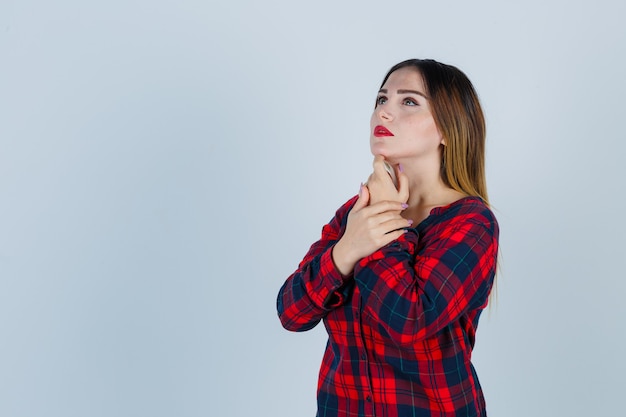 Young woman propping chin on finger, looking up in checked shirt and looking pensive , front view.