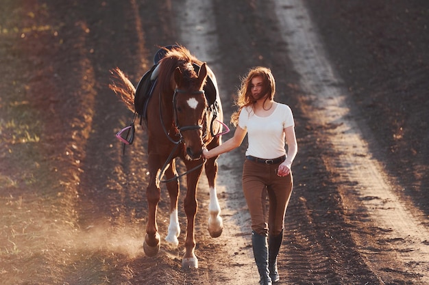 Young woman in protective hat walking with her horse in agriculture field at sunny daytime