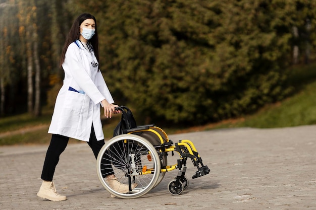 Young woman in a protective mask and medical gown carries a wheelchair Attractive nurse looks at the camera carries a wheelchair outdoors