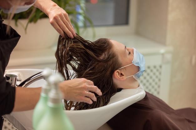 Young woman in protective medical mask getting a hair wash in beauty salon during pandemic