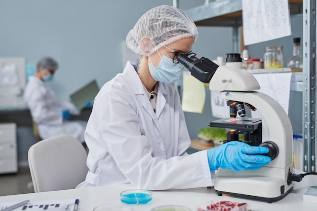 Young woman in protective workwear sitting at the table and examining samples with microscope at the lab