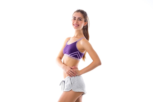 Young woman in purple sports top and shorts showing her abdominals on white background in studio