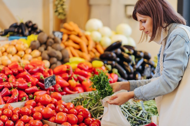 Young Woman puts fruits and vegetables in cotton produce bag at food market. Reusable eco bag for shopping. Sustainable lifestyle. Eco friendly concept.