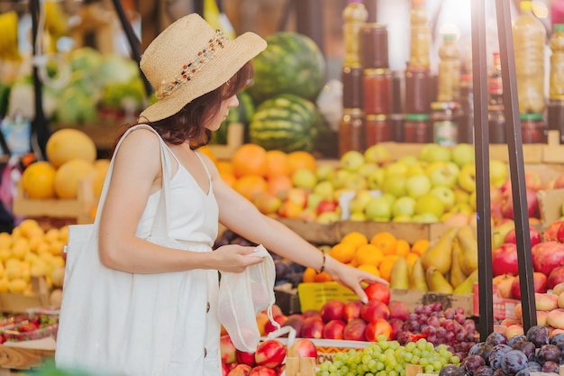 Young Woman puts fruits and vegetables in cotton produce bag at food market. Reusable eco bag for shopping. Zero waste concept.