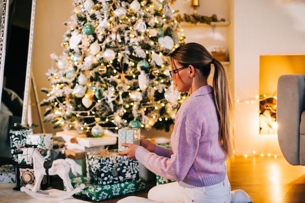 Young woman puts present under Christmas tree while sitting on the floor at home.