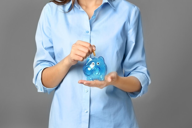 Young woman putting coin into piggy bank on grey background