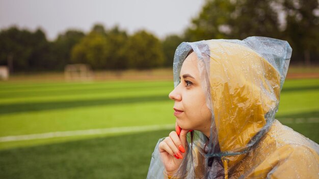 Young woman in raincoat sitting on football field in rainy weather Positive female in wet waterproof coat enjoying weather