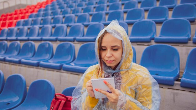 Young woman in raincoat with notepad pen sitting on stadium bleachers alone in rainy weather Female journalist writing down notes during sports training at street stadium