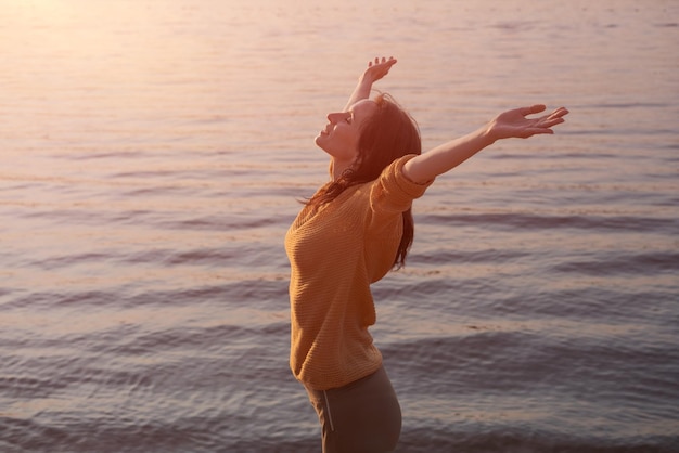 Young woman raises her hands to setting sun on shore lake in yellow sweater