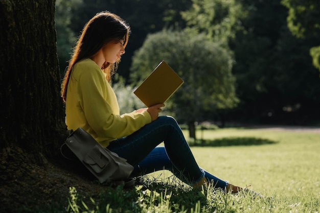 Young woman reading book near tree in park on sunny day