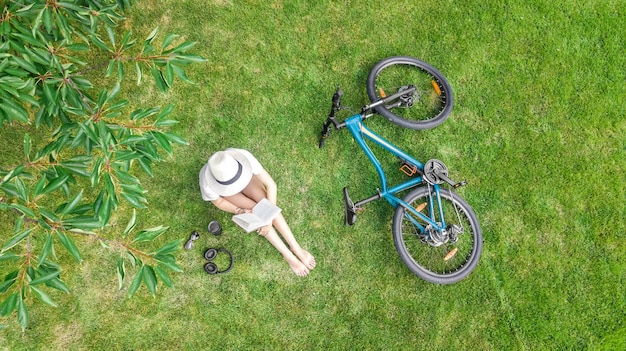 Young woman reading book in park near bike. Student girl relaxing outdoors sitting on grass