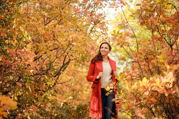 Young woman in red coat walking in sunny autumn park with yellow leaves