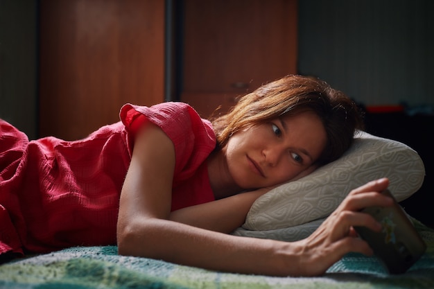 Young woman in red dress lying on the bed and watching video on smartphone