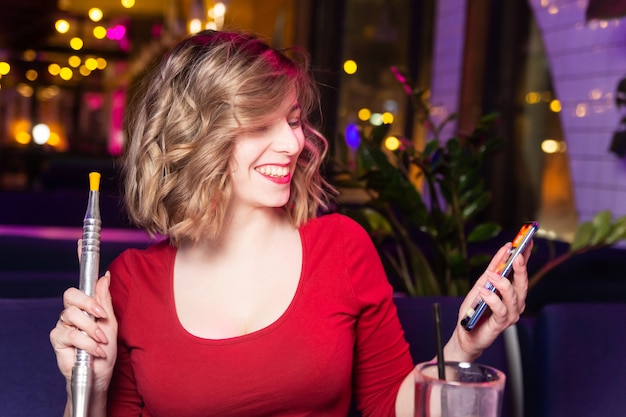 Young woman in the red dress smokes a hookah at the hookah bar and chatting with friends. 