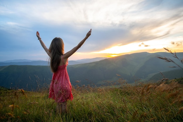 Young woman in red dress standing on grassy field on a windy evening in autumn mountains raising up her hands enjoying view of nature