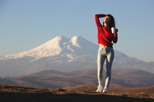 Young woman in a red sweater standing against the blurred mountains enjoying the first rays of the sun with cup of tea in her hand