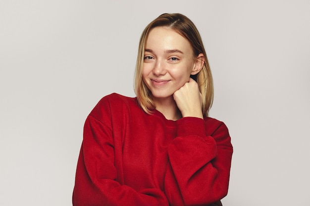 Young woman in red sweatshirt smiles positively holds chin with both hands