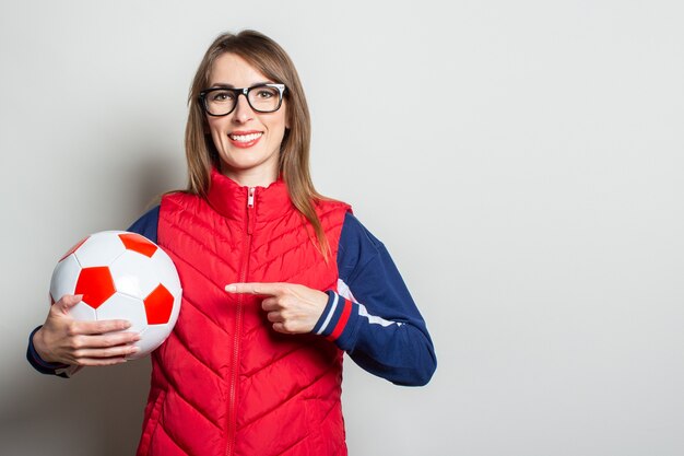 Young woman in a red vest holds a soccer ball in her hands and points at it with her finger