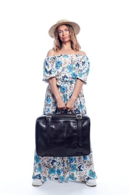 A young woman in a romantic summer dress and a straw hat is holding a heavy retro leather suitcase Leisure vacation and travel Isolated on white background Vertical