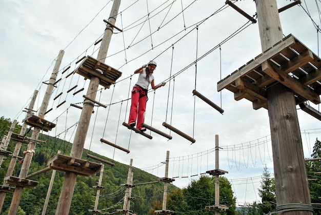 A young woman in a rope amusement park crosses a suspension wooden bridge in a harness