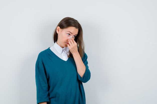 Young woman rubbing nose, looking away in sweater over white shirt and looking thoughtful , front view.