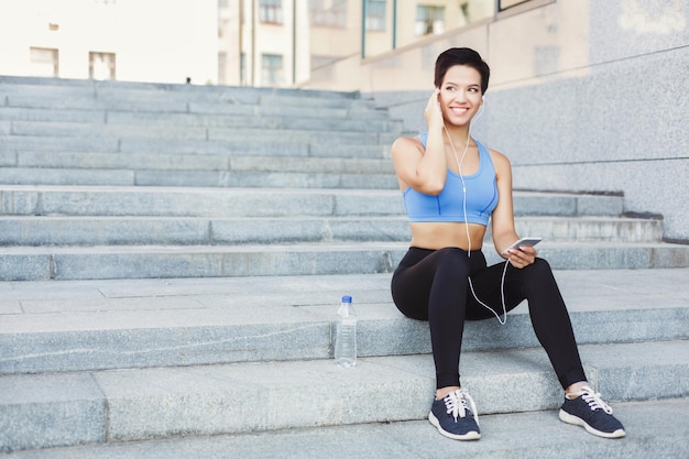 Young woman runner is having break, enjoying music in headphones and drinking water while jogging in city, sitting on staircase and smiling, copy space