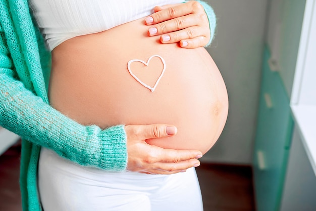 Young woman's hand holding a white heart shape on a naked belly. Close-up.