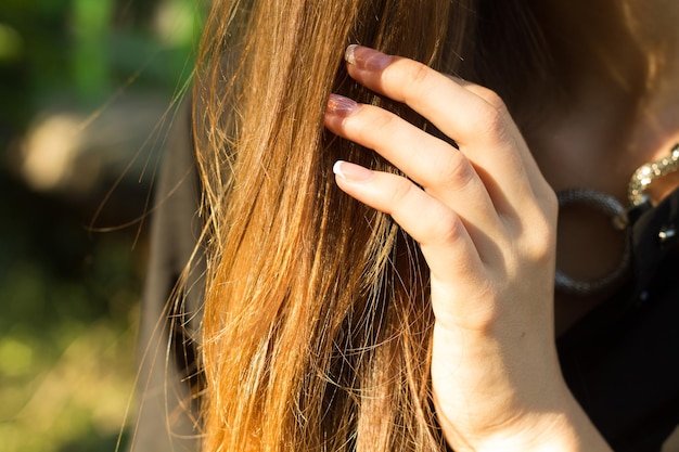 Young woman's hand touching her long brown hair on the background of green trees