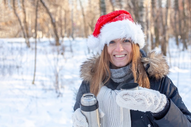 A young woman in a santa claus hat pours herself some hot tea from a thermos.