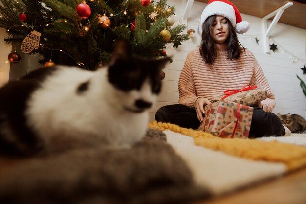 Young woman in santa hat opening christmas gift with her cat under christmas tree in festive room