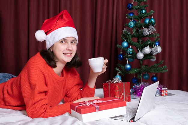 A young woman in a Santa hat and a red sweater holds a mug of coffee or tea, rests with her laptop, lying on the bed, surrounded by Christmas gifts. Christmas online shopping