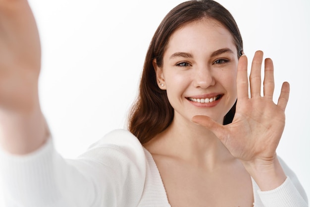 Young woman saying hello via video call holding smartphone and taking selfie as waving hand and smiling friendly standing over white background