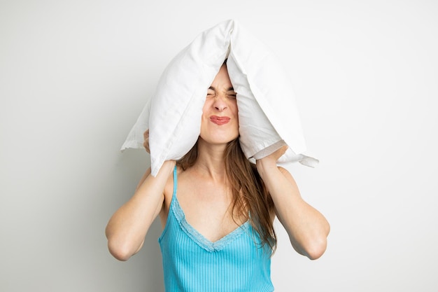 Young woman screwing up her eyes closed her ears with a pillow on a white background