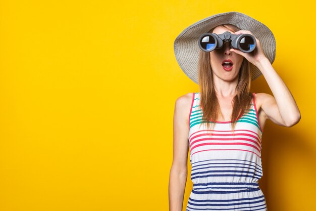 Young woman in shock in surprise with a hat and a striped dress looks in surprise with binoculars on a yellow space