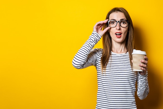 Young woman shocked in glasses with a paper cup of coffee on a yellow background