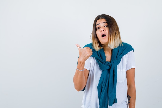 Young woman showing thumb up in white t-shirt and looking surprised , front view.