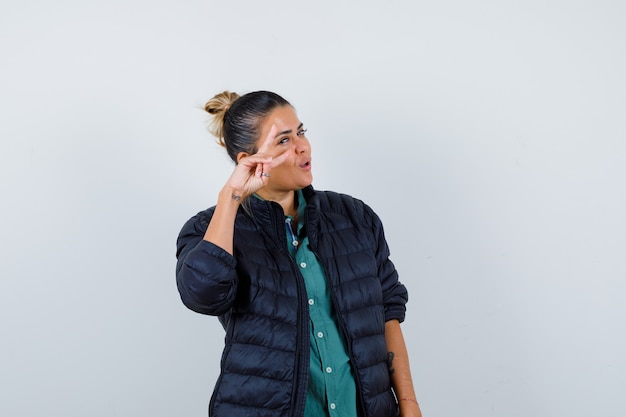 Young woman showing V-sign near eye in shirt, puffer jacket and looking lucky. front view.