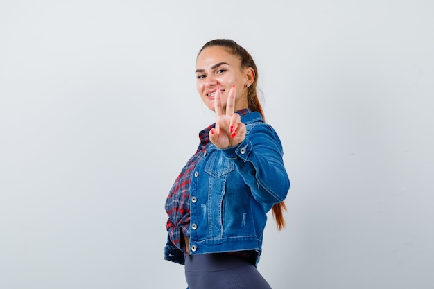 Young woman showing V-sign, standing sideways in checkered shirt, jean jacket and looking attractive .