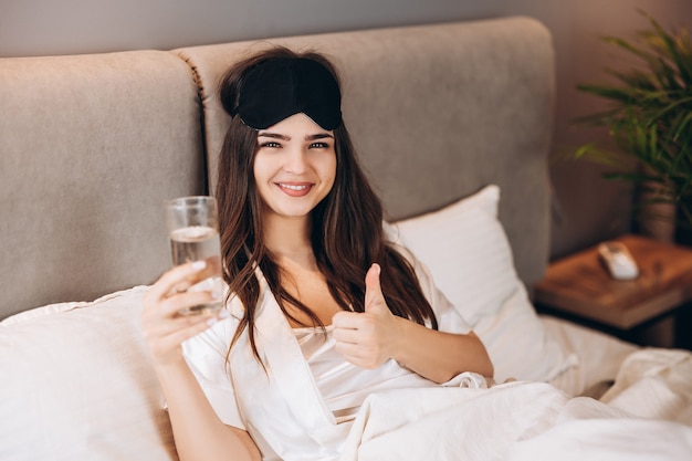 Young woman shows that drinking water is in the bed. model with brown hair with a glass of water in the bed in the bedroom. drink a glass of water in the morning or before bed.