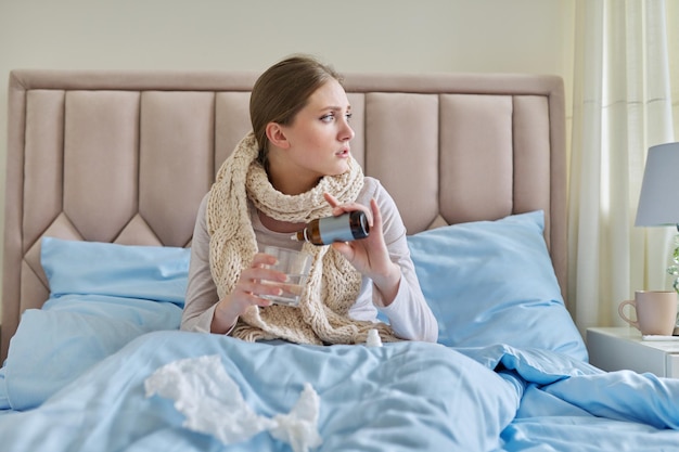 Young woman sick at home in bed female holding glass of water and dripping medicine