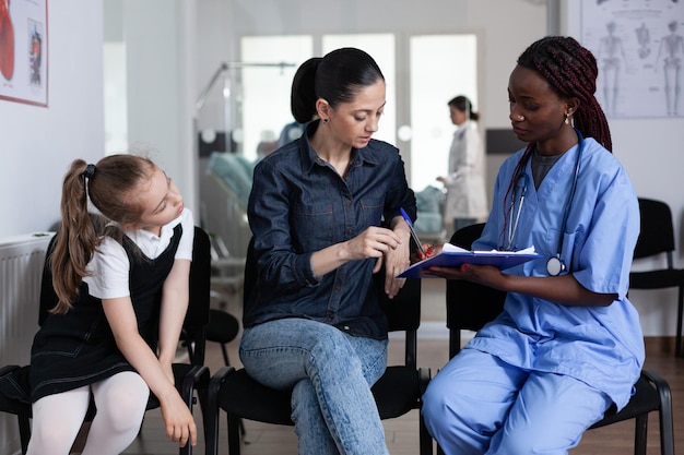 Young woman signing little girl discharge papers in children hospital waiting room. Mother filling out daughter file with african american pediatrician at medical clinic lobby.