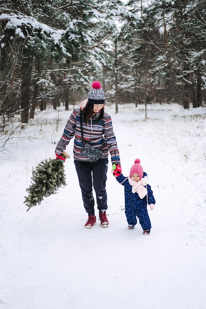 Young woman single mom and toddler baby girl carries christmas tree in the winter park preparing for