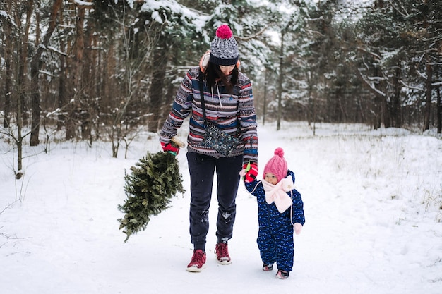 Young woman single mom and toddler baby girl carries christmas tree in the winter park preparing for