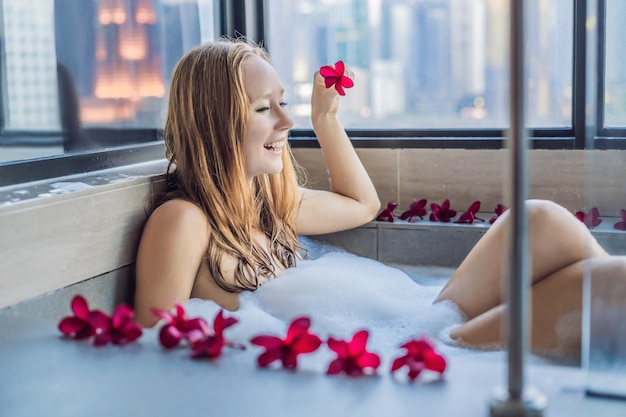 Young woman sits in a bath with foam and frangipani flowers against the background of a panoramic window overlooking the skyscrapers and a big evening city