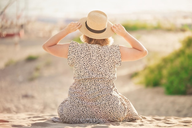 Young woman sits on the beach sand holds a hat with her hands and looks into the distance towards